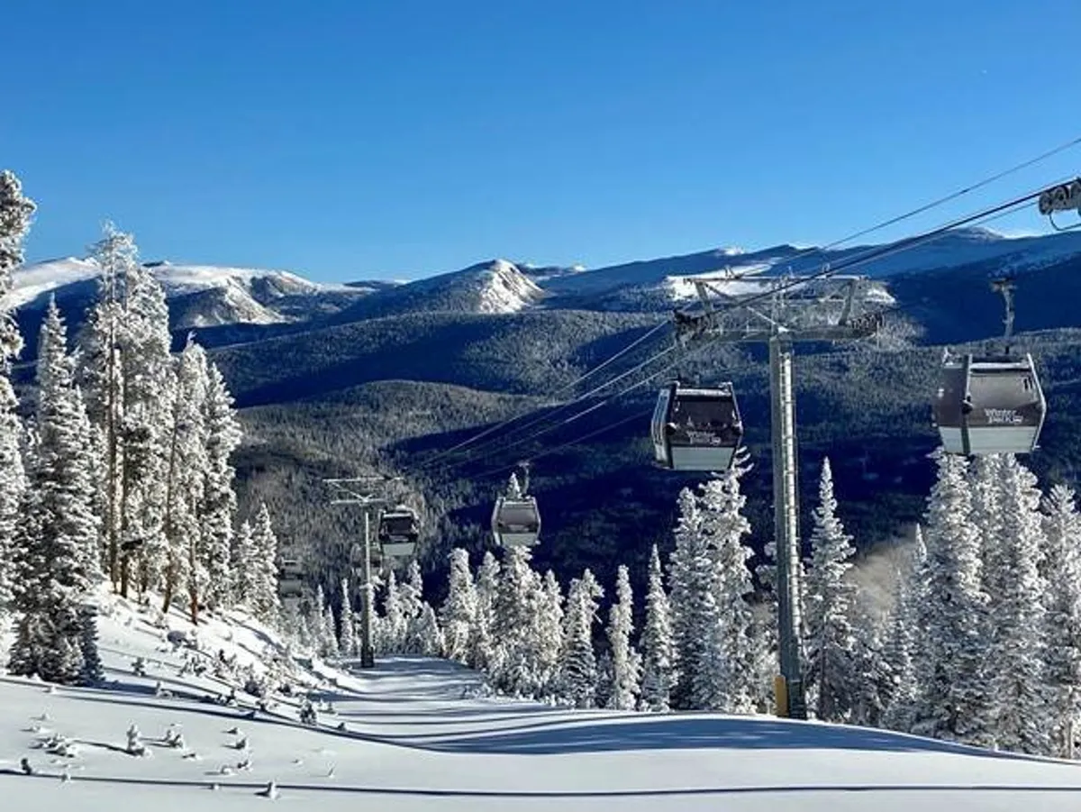 A stunning view of snow-covered mountain slopes with a ski lift ascending through evergreen trees, set against a dramatic mountain range. The scene captures the excitement and beauty of Winter Park’s ski and snowboard offerings in peak winter.