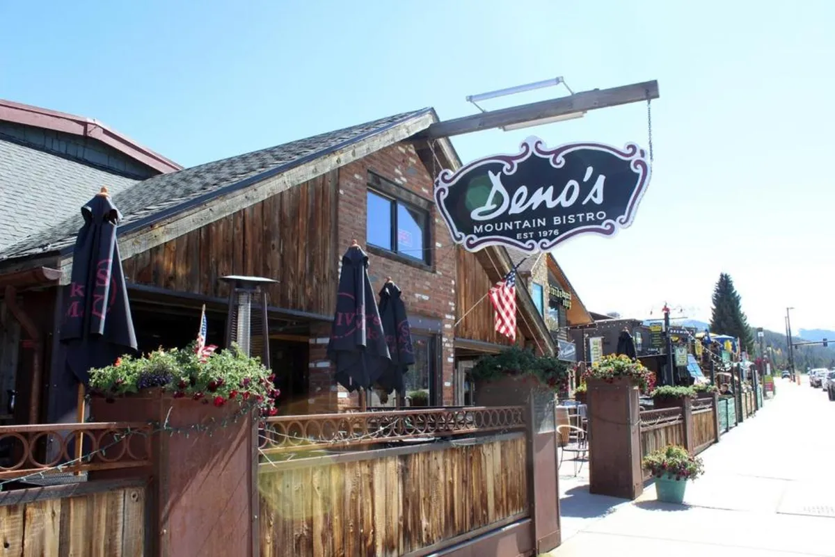 The photo highlights Deno's Mountain Bistro's warm wooden facade, outdoor seating surrounded by flowerpots, and guests enjoying meals al fresco. The inviting scene blends mountain charm with the vibrant social life of downtown.