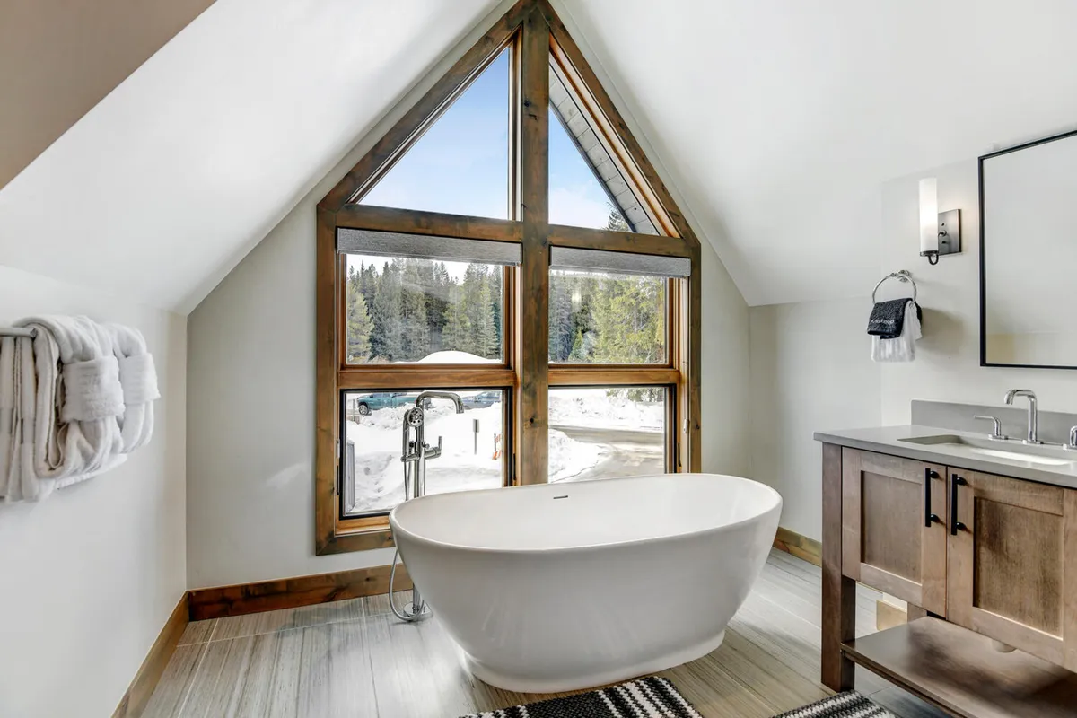 A spa-inspired bathroom with a freestanding tub positioned by a large triangular window overlooking snowy mountains. Wood cabinetry and natural light enhance the sense of tranquility and luxury.