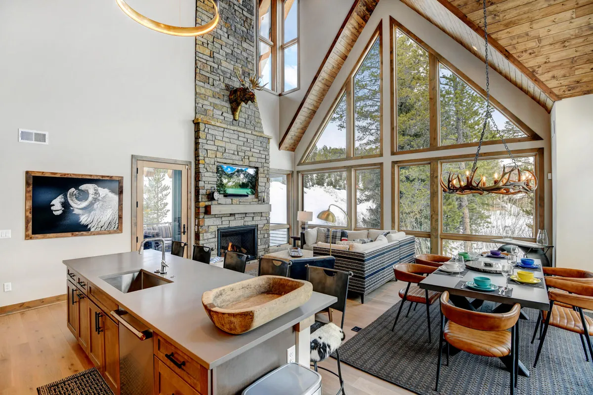 The image showcases a spacious, light-filled living and dining area featuring soaring windows framing scenic mountain views, a striking stone fireplace, a modern dining table set for gatherings, and a rustic kitchen island—combining luxury and warmth. Natural light bathes the space, inviting guests to relax, dine, and connect in style.