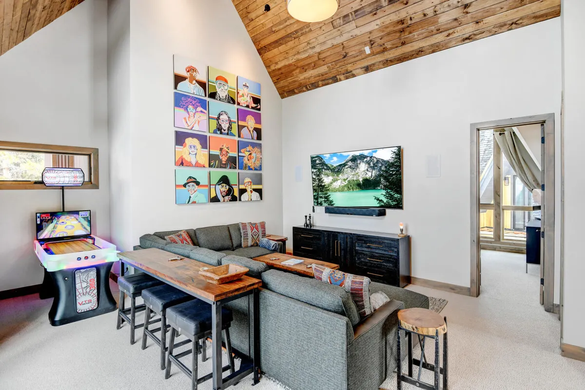 A lively rec room featuring a colorful wall of portraits, a modern flat-screen TV, a cozy sectional sofa, an arcade game machine, and a rustic wooden table. The space is set up for movie nights, game tournaments, and group gatherings with easy access to snacks and drinks.