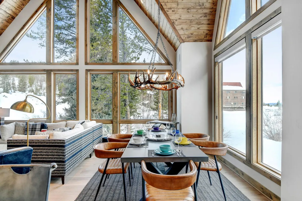 An open interior scene showcasing a spacious dining area set for a meal, comfortable striped sofa, and large windows offering expansive mountain views. An antler chandelier hangs above the concrete dining table, with the overall atmosphere exuding warmth and togetherness.