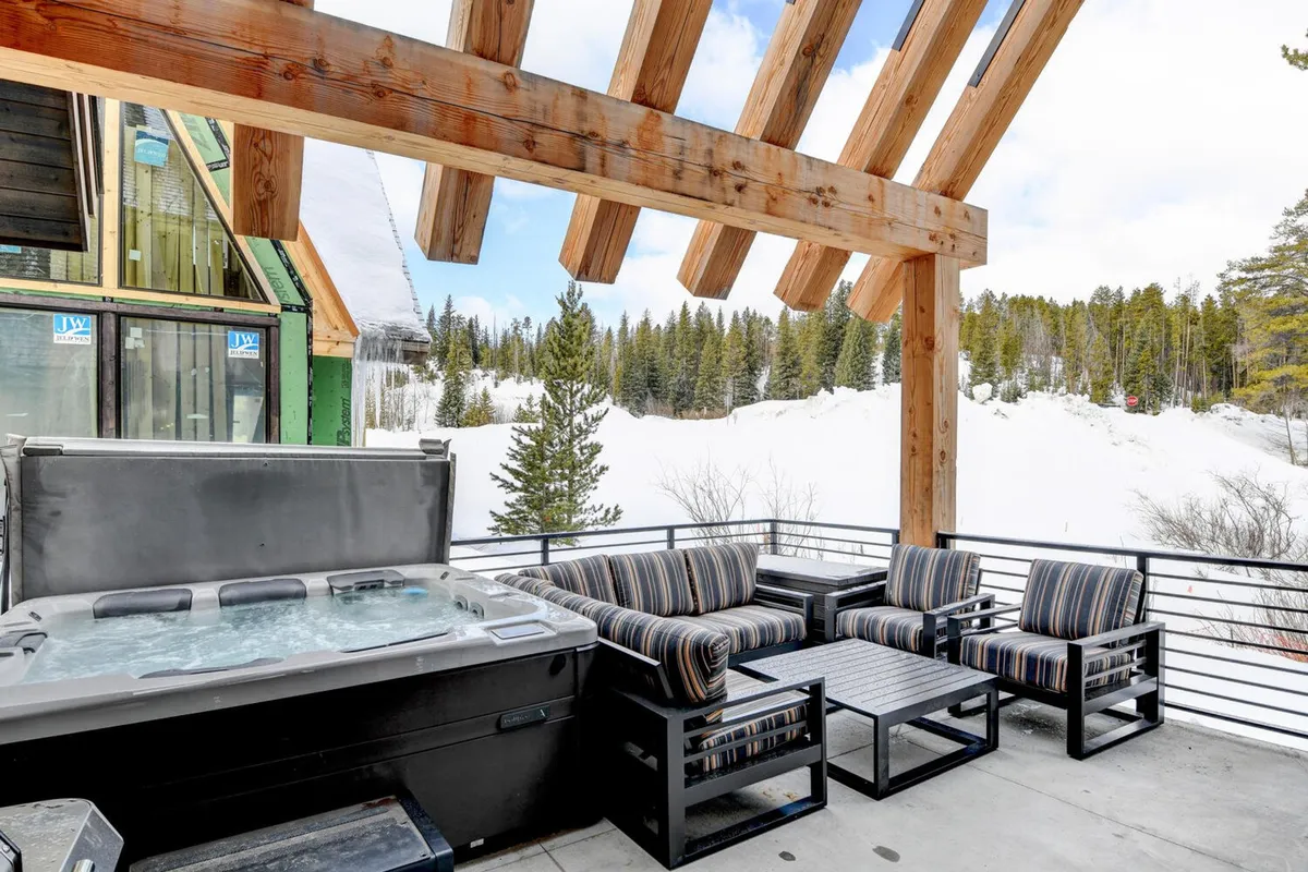 An inviting exterior shot of a chalet's outdoor hot tub area, surrounded by a snowy landscape. The scene features striped seating, wooden beams, and the bubbling hot tub, evoking relaxation and comfort.