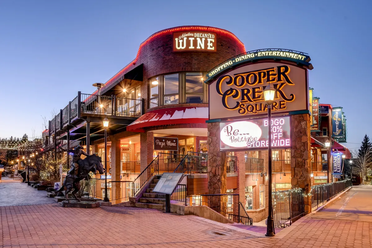 This image showcases the lively exterior of Cooper Creek Square, featuring signage for dining and wine shops, outdoor seating areas, and festive decorative lighting. People can be seen enjoying the vibrant atmosphere, highlighting a hub of social activity at the heart of downtown.