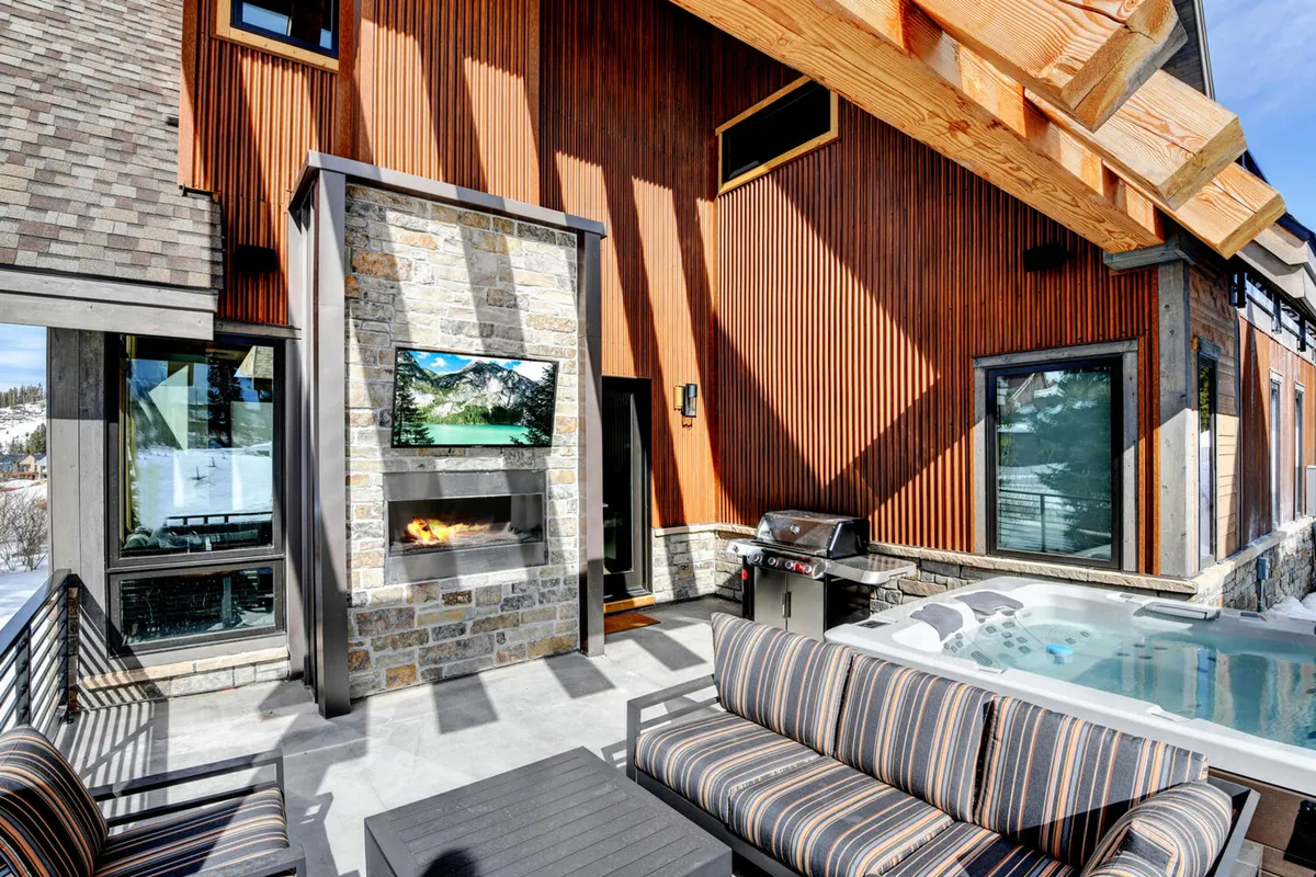 This exterior scene showcases a stone fireplace, an outdoor television, a bubbling hot tub, and comfortable outdoor seating on a patio surrounded by mountain views. The atmosphere is relaxed and luxurious, with elements that invite guests to socialize, barbecue, watch TV outside, or unwind in the hot tub while taking in the sunset.