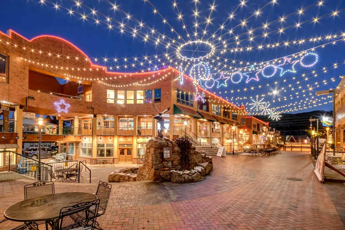 A charming downtown scene illuminated by twinkling overhead lights, decorative stars, and a stone fountain. Outdoor seating invites social gatherings, and the festive mood suggests a lively community hub year-round.