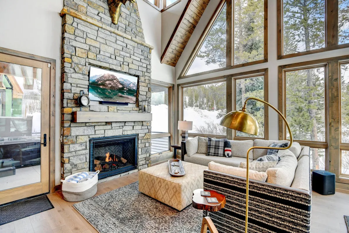 A cozy living area with a custom stone fireplace, plush sofa, large windows framing snowy views, and tasteful wall art. The setting is ideal for relaxing, socializing, or gathering for movie nights.